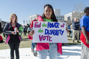 A young woman in a dispersed crowd hold a sign stating "Feel the power of the disability vote"