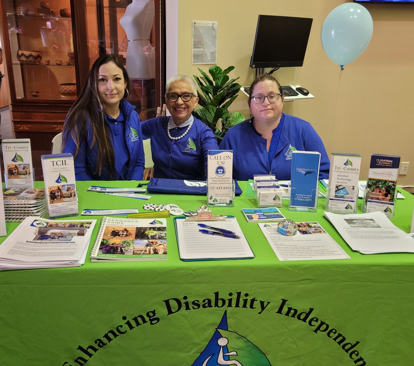 ADRC Program Coordinator Lisa Leon, ILS/Program Coordinator Mari Dorenstreich, Assistive Technology Coordinator Juliannah Haris wearing their TCIL blue branded jackets are seated at a booth with the "Tri-County Independent Living" banner, showcasing various informational materials and promotional items related to disability services.