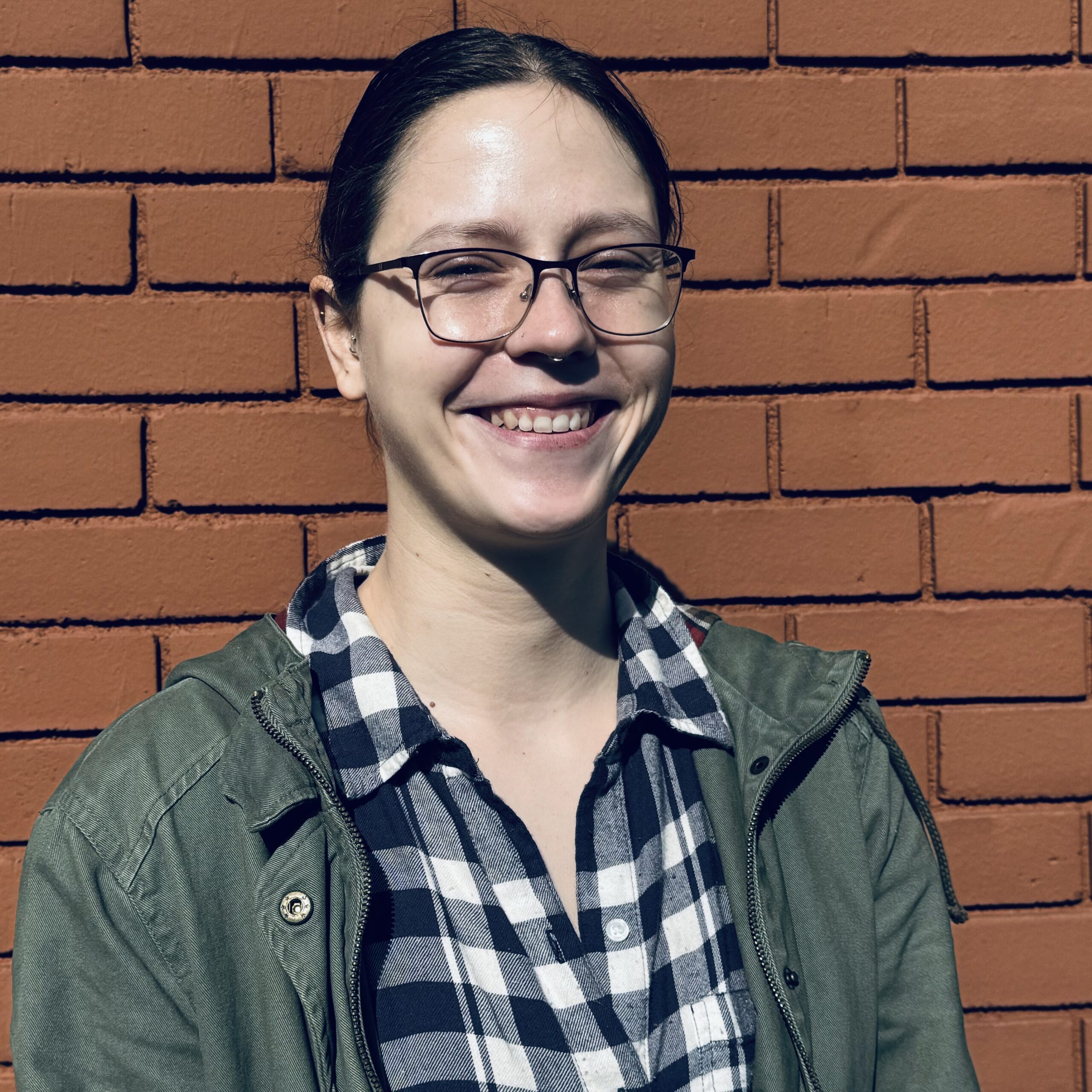 Denise a female with glasses and a plaid shirt smiles while standing in front of an red brick wall.