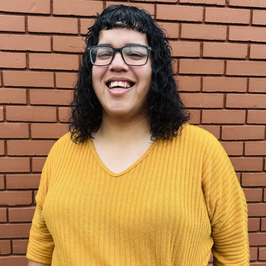 Photo of Anisa Escobedo wearing a yellow sweatshirt, black hair, with black glasses.